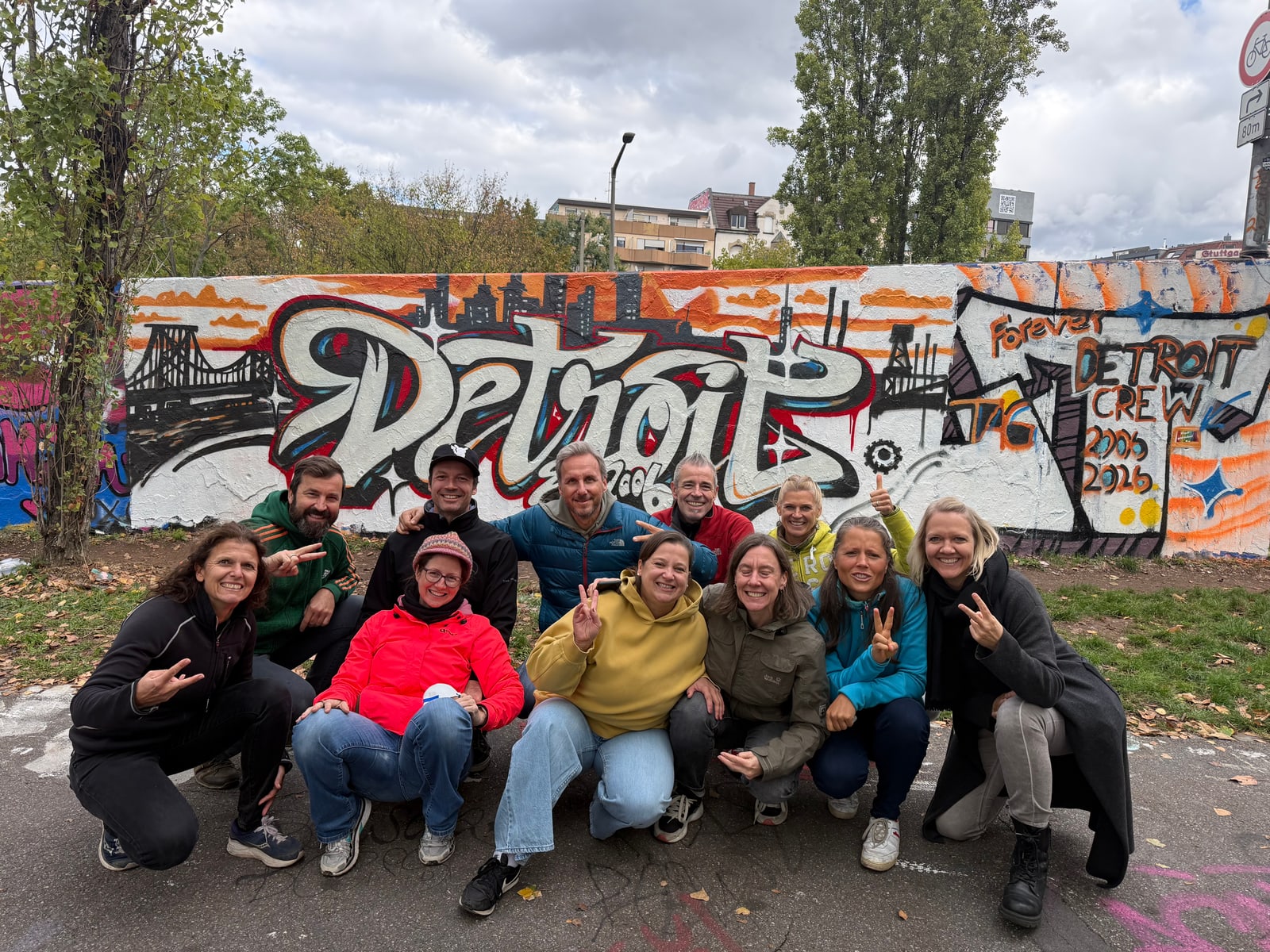 Zwölf Freundinnen und Freunde hocken und stehen lachend vor der fertigen Graffiti-Wand mit dem Schriftzug Detroit und dem Feld Forever Detroit Crew.