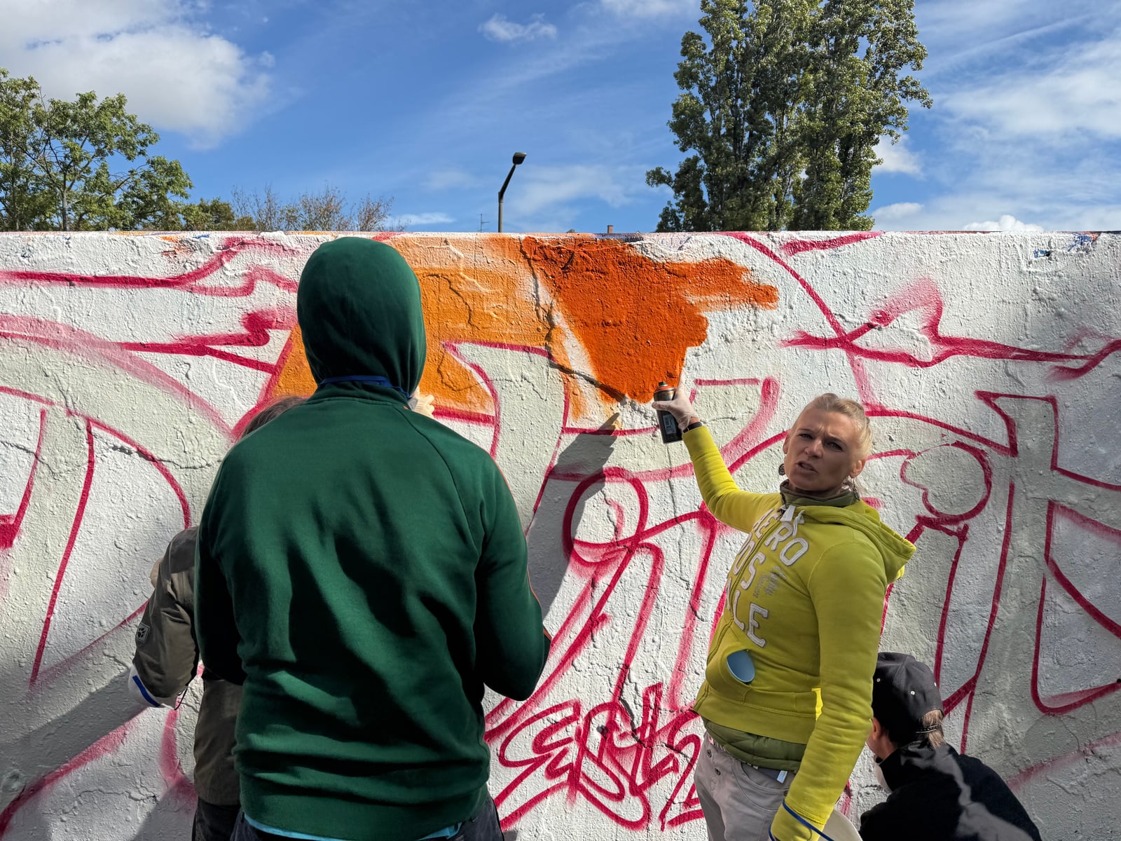 Eine Teilnehmerin in gelber Jacke sprüht die erste orange Fläche auf die weiße Wand, auf der die Skizze in Rot vorgezeichnet ist.