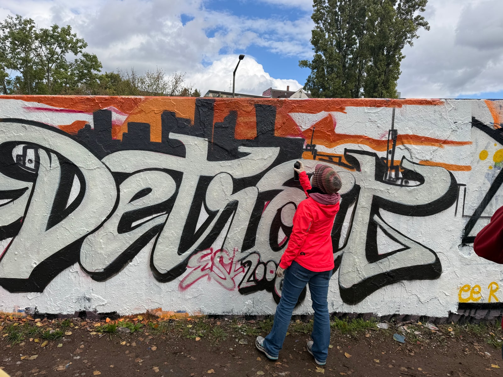 Eine Teilnehmerin in roter Jacke sprüht am großen schwarz-weißen Schriftzug Detroit vor oranger Skyline.
