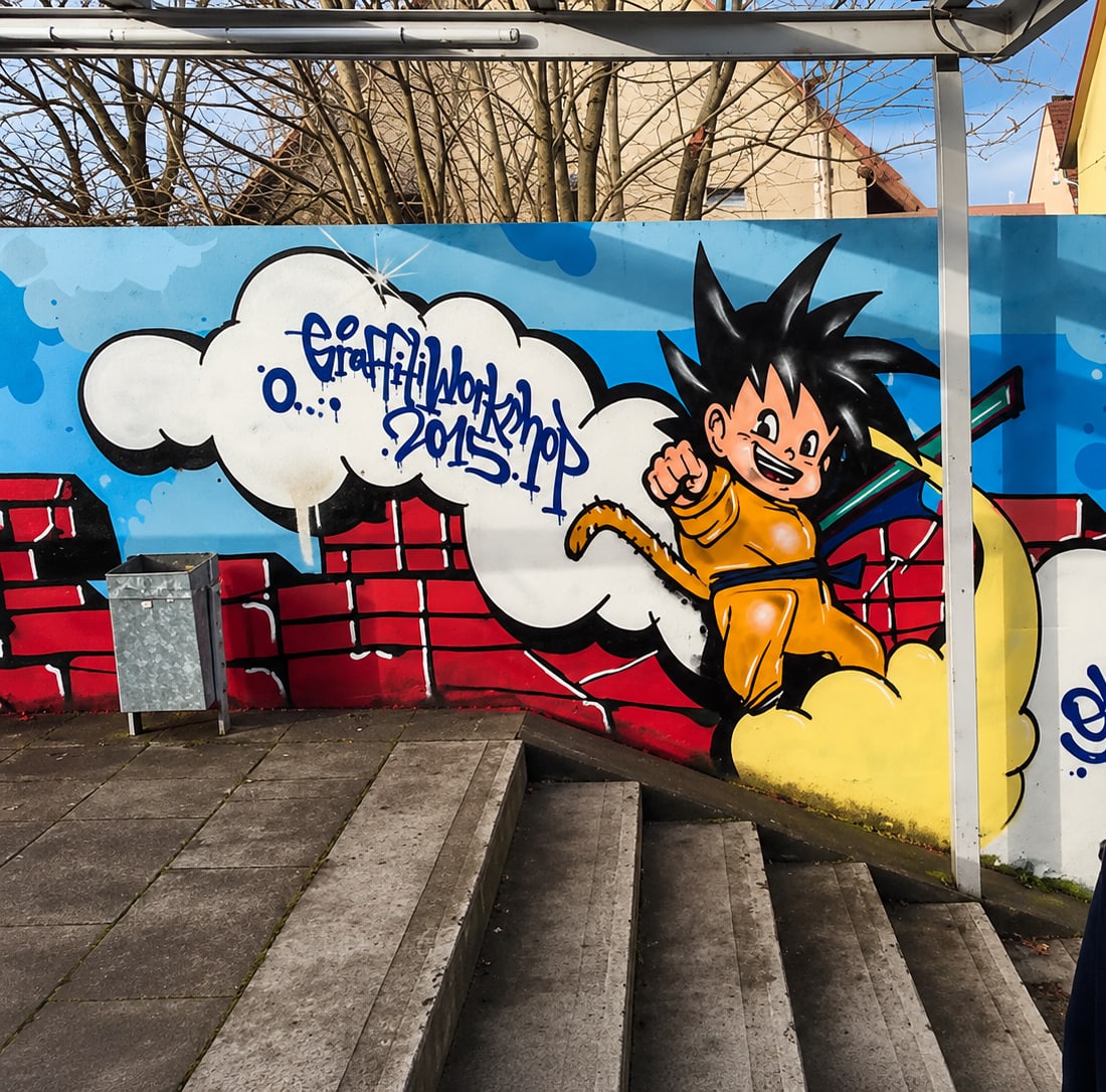 Gemalte Manga-Figur mit schwarzen Stachelhaaren im orangefarbenen Kampfanzug springt aus einer weißen Wolke, dahinter blauer Himmel und eine gemalte rote Backsteinmauer.