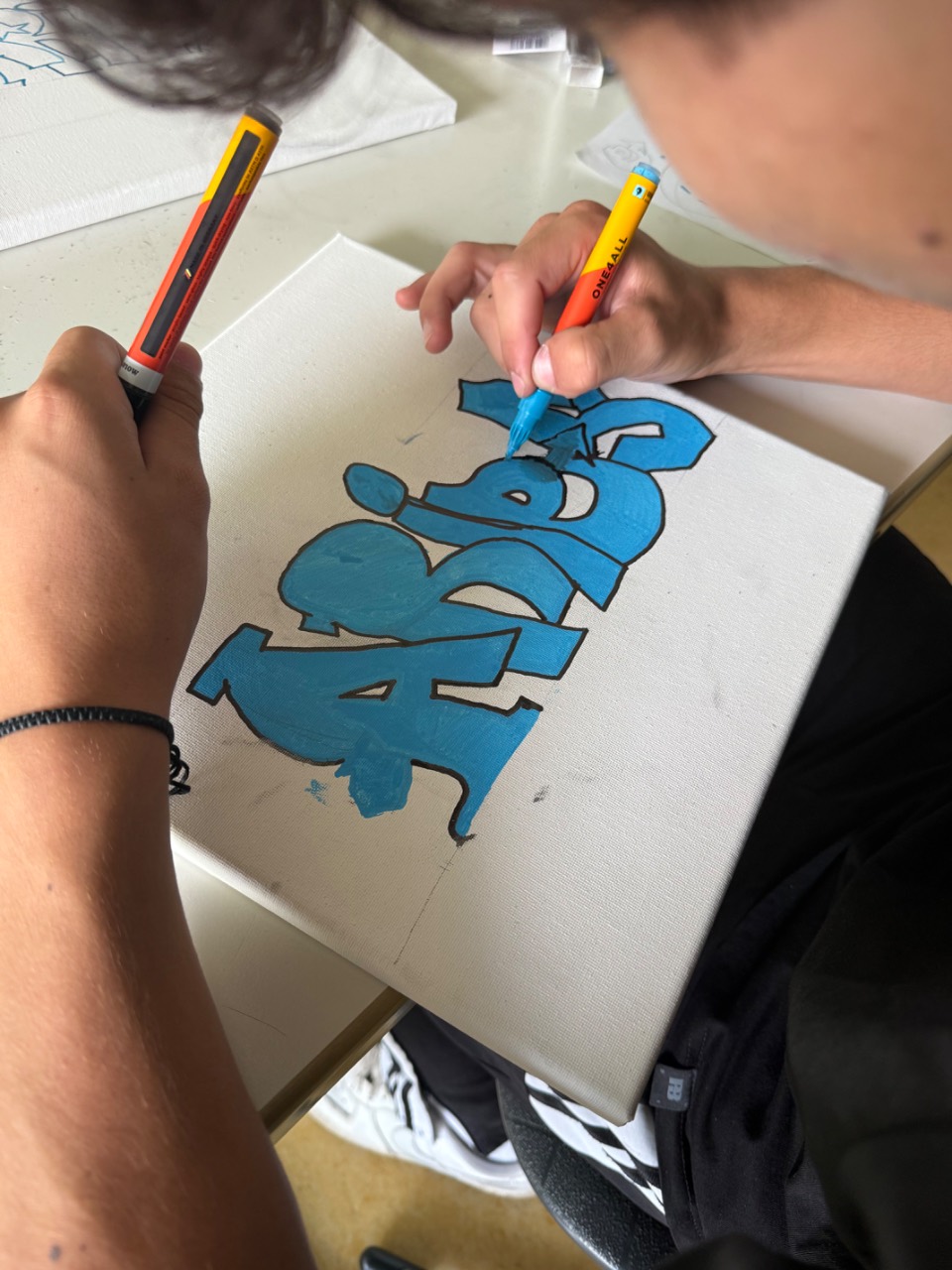 Zwei Personen malen gemeinsam mit blauen Markern einen dicken Graffiti-Schriftzug auf eine weiße Leinwand, Blick von oben auf Hände und Leinwand.