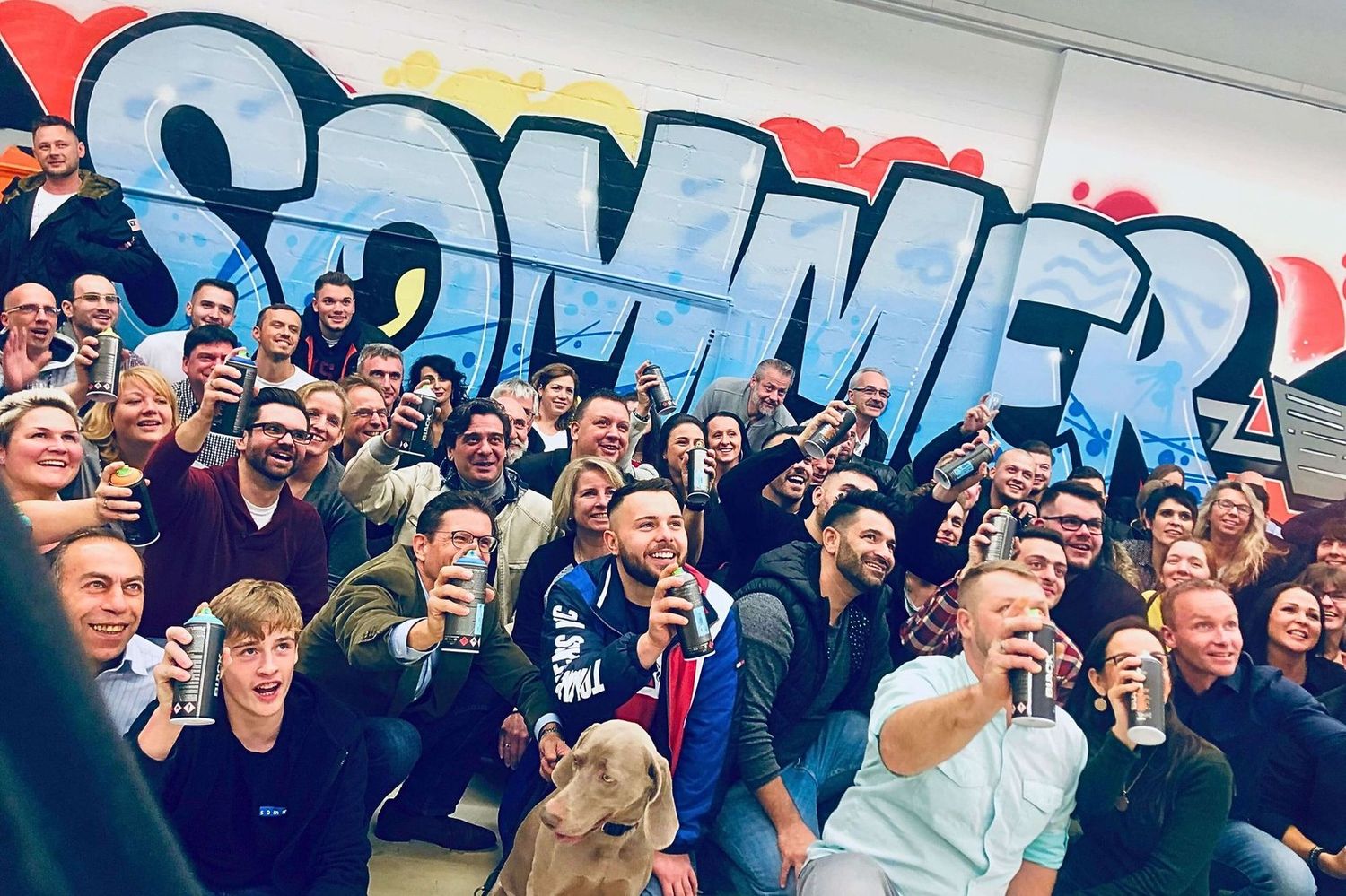 Teilnehmer des Graffiti Teambuilding Workshops bei der Firma Sommer.de posieren vor dem gemeinsam gesprühten Graffiti.