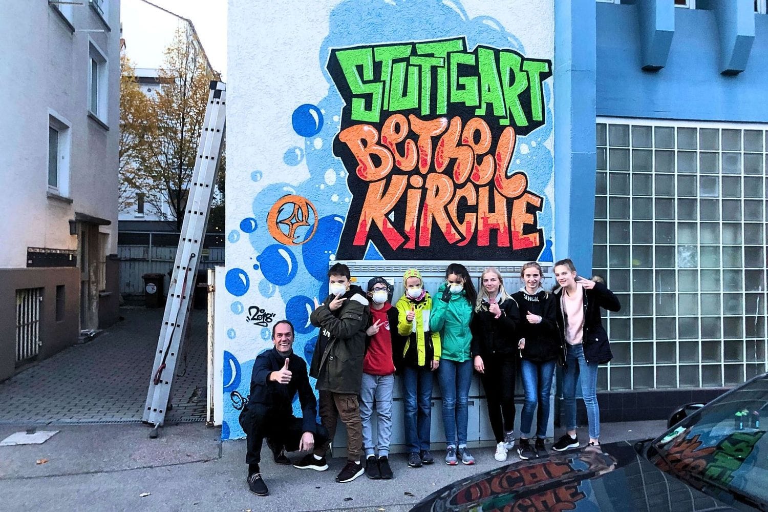 Gruppenbild mit Jugendlichen und Open Mike vor dem fertig gesprayten Graffiti „Bethel Kirche Stuttgart“ – Urban Art Workshop an der Kirchengemeinde.