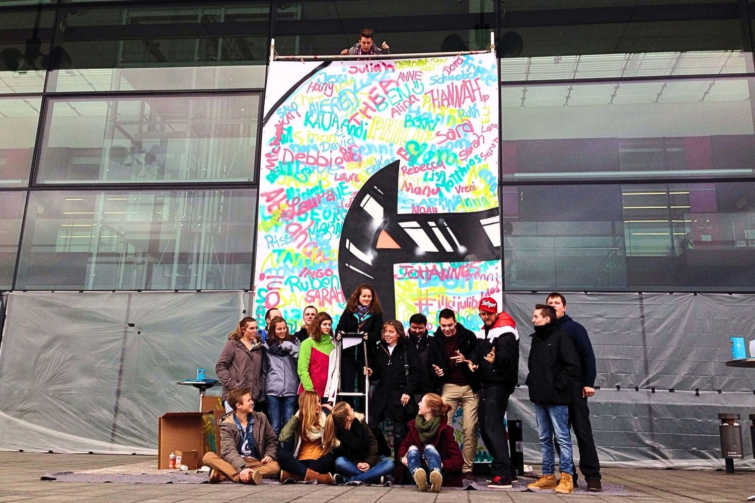 Open Mike und Teilnehmende posieren vor dem fertig gestalteten „C“ des Christus-Banners beim Vor-Workshop am Flughafen Stuttgart.