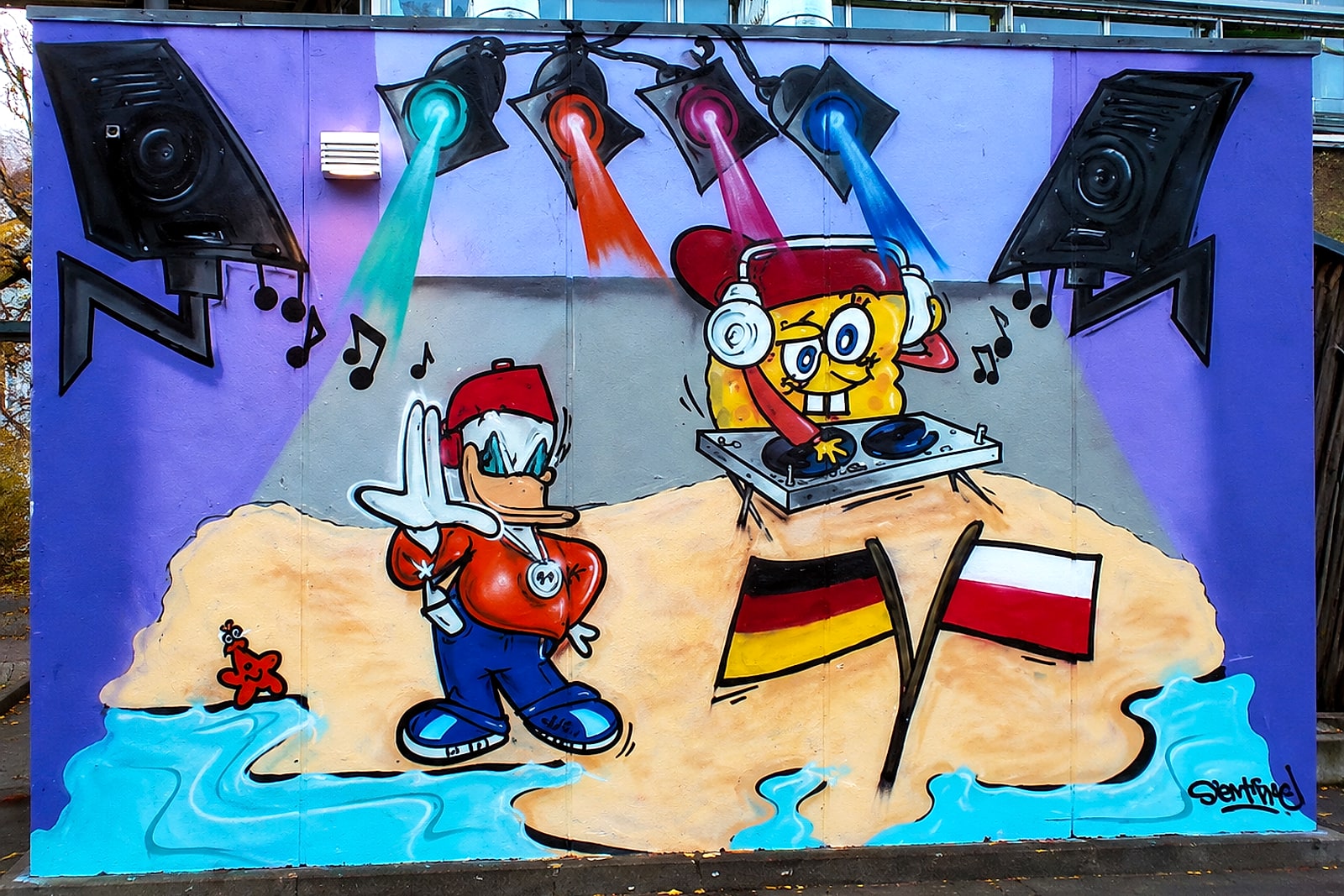 Comic-Party-Szene der Fassade am Jugendhaus Hausen: DJ-Figur, Boxen, Scheinwerfer, Deutschland- und Polen-Flagge, Strand und Wasser.