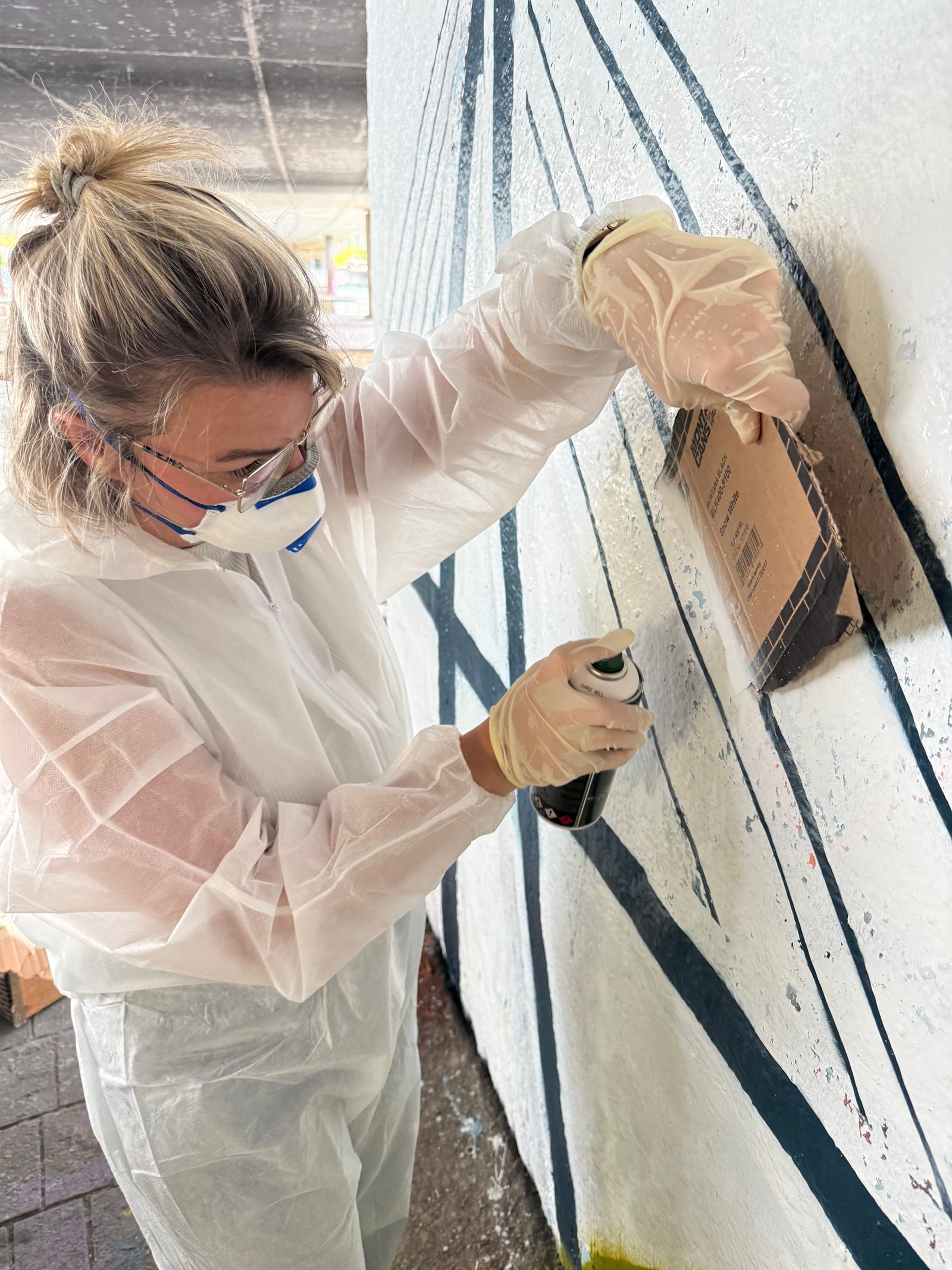 Frau in weißem Overall, Brille und Atemmaske sprüht mit Dose gegen eine als Schablone gehaltene Pappe an der Wand.
