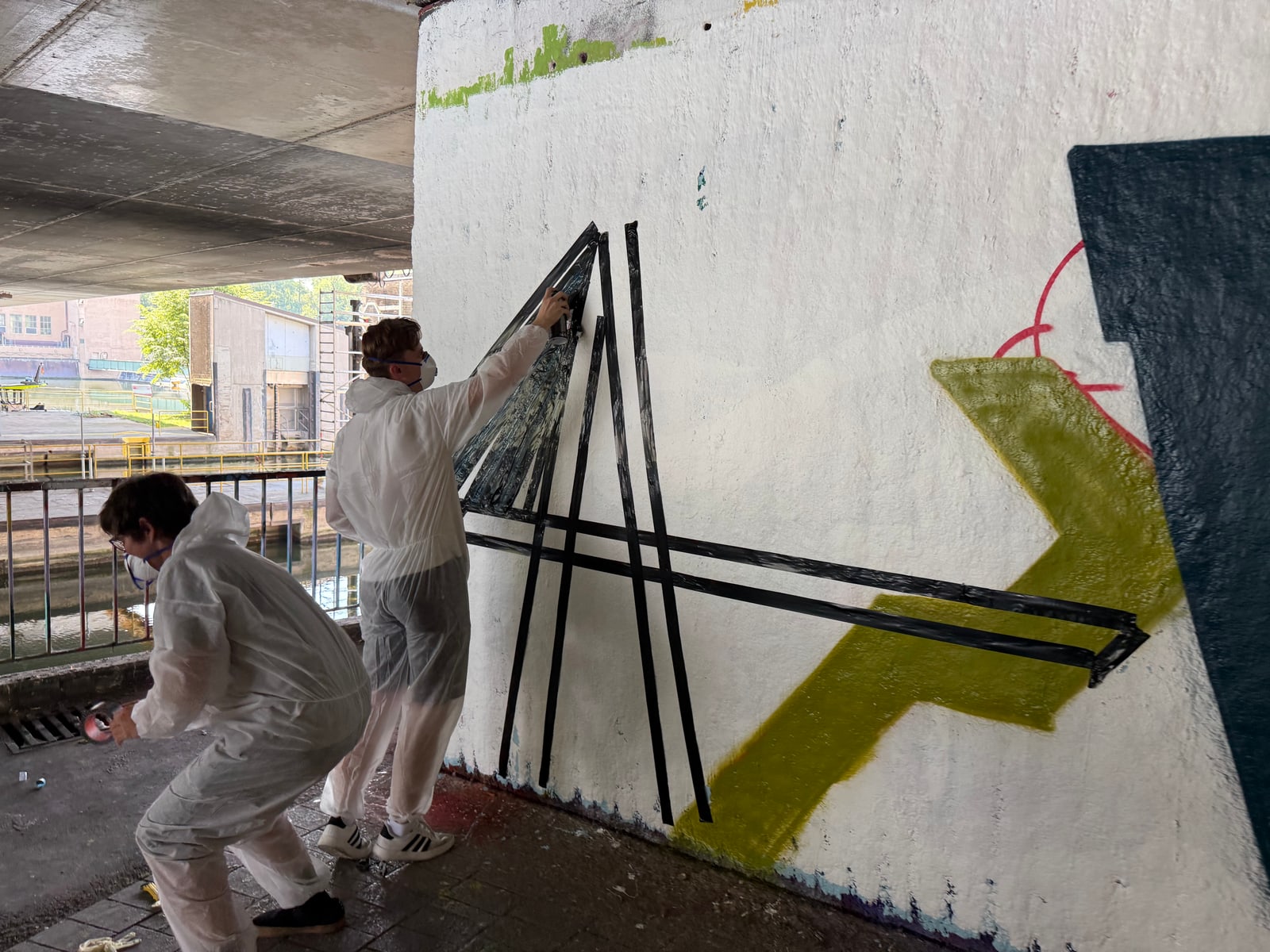 Zwei Personen in weißen Overalls und Atemmasken kleben schwarze Klebebandlinien auf eine weiße Wand unter der Brücke.