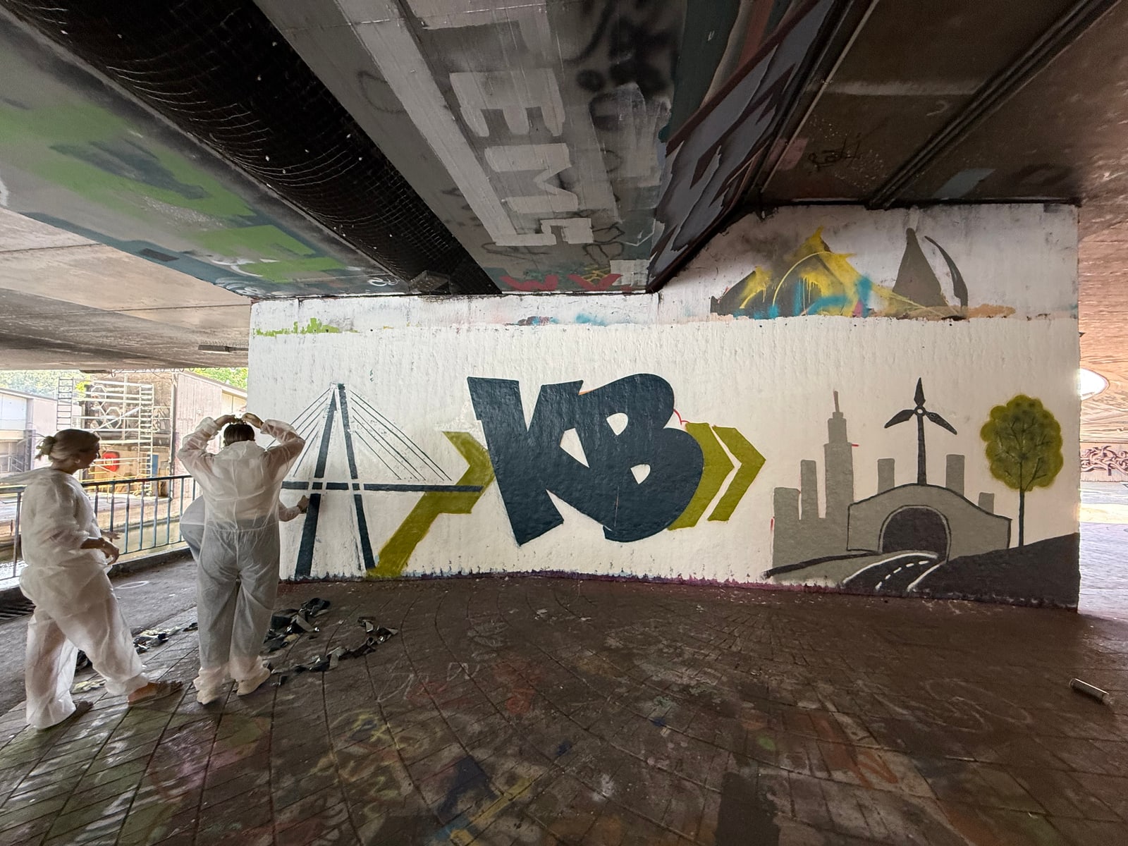 Zwei Personen in weißen Overalls vor der Wand mit KB-Schriftzug, Brückenmotiv, Skyline und Baum, Klebeband am Boden.