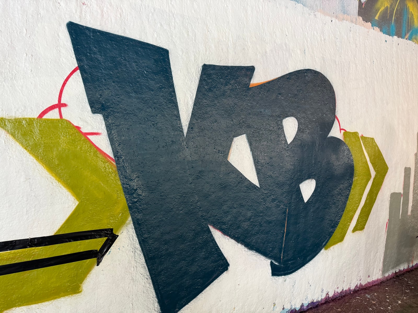 Nahaufnahme der dunkelblauen Buchstaben K und B auf weißer Betonwand, olivgrüner Pfeil und rote Umrisslinie.