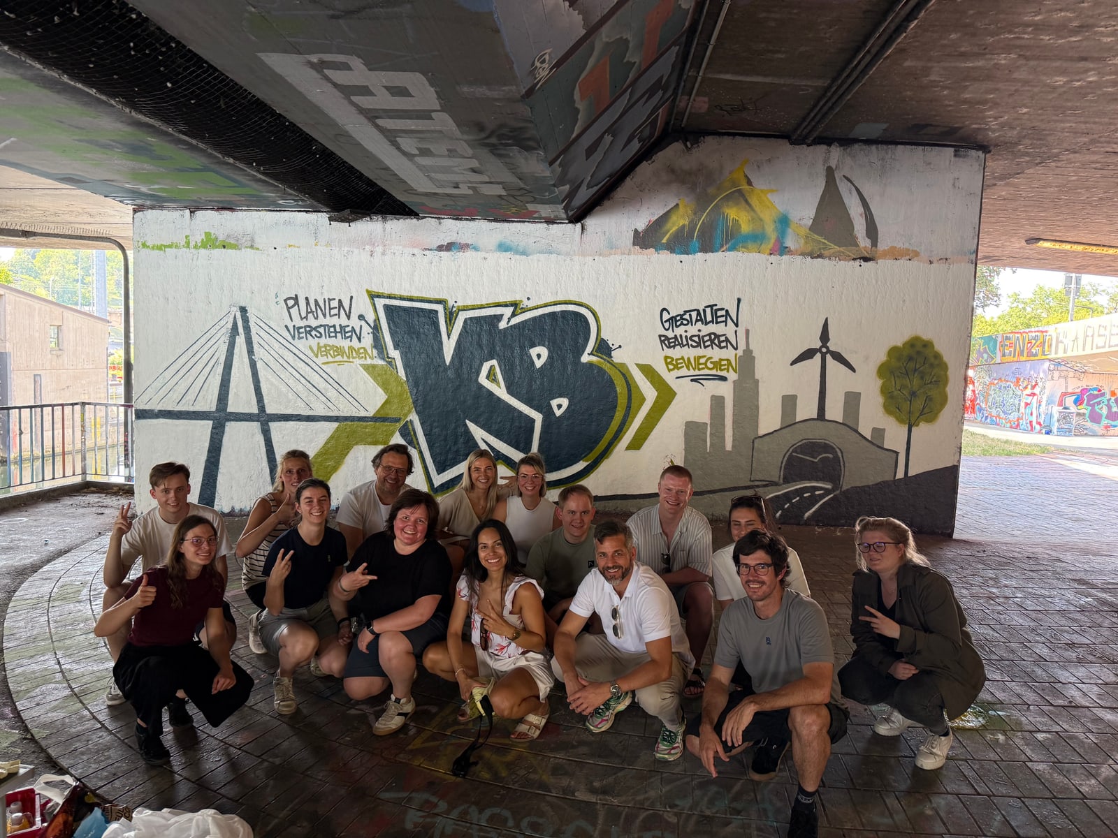 Gruppe von etwa 17 Personen posiert vor fertigem Wandbild mit KB-Schriftzug, Brücke, Skyline und Baum.