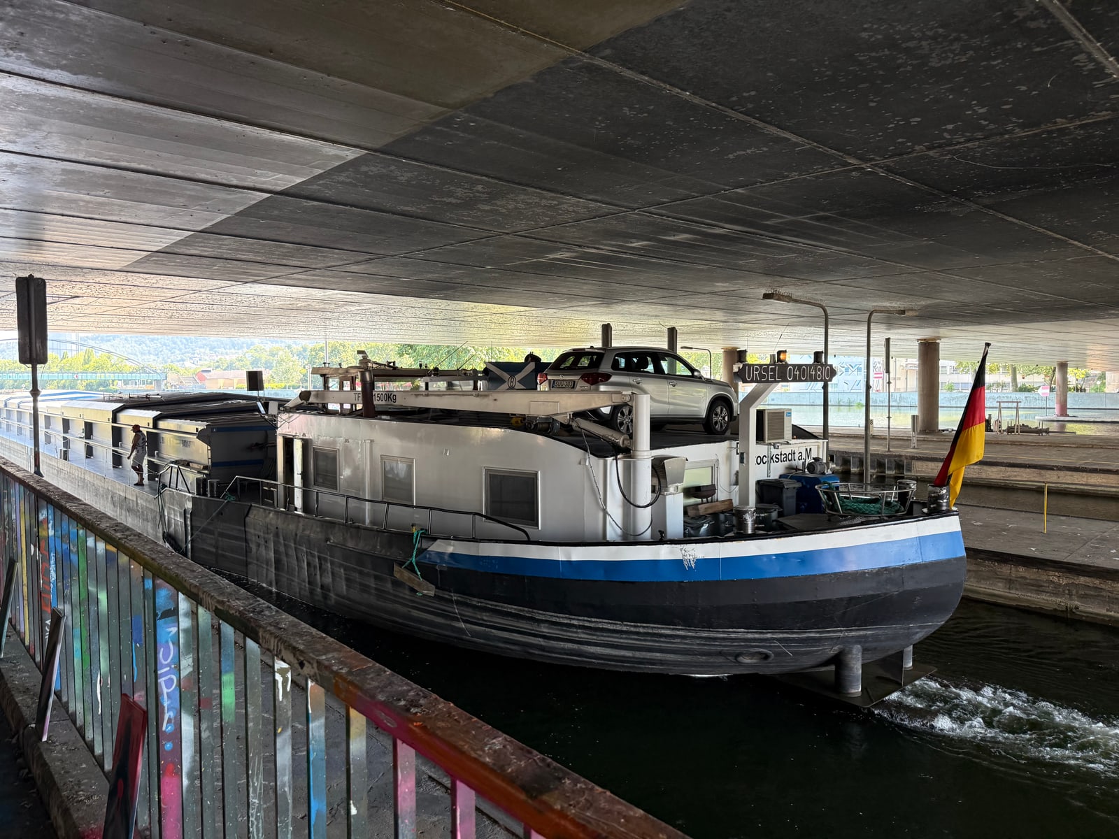 Frachtschiff mit Auto an Deck fährt unter einer Betonbrücke durch, deutsche Flagge am Heck, buntes Geländer links.
