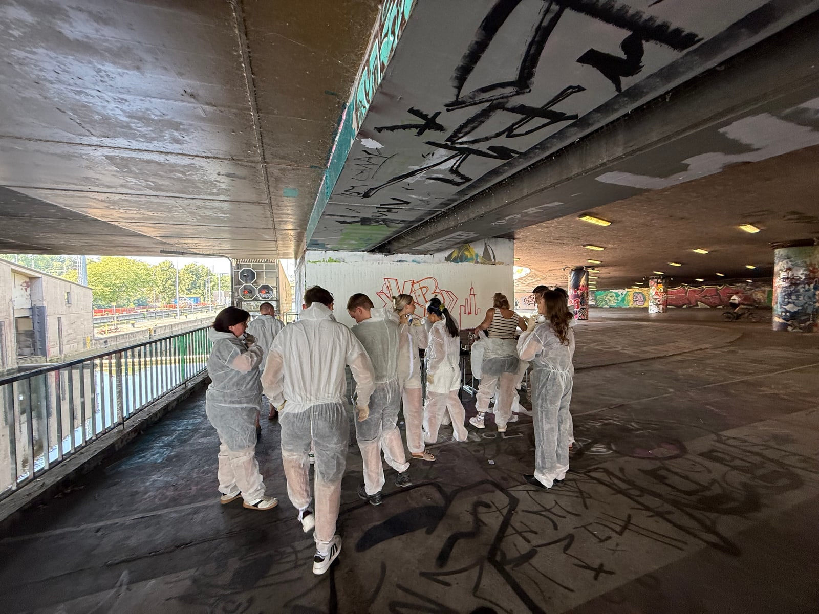 Gruppe in weißen Einwegoveralls steht unter einer Betonbrücke vor weiß grundierter Wand, Fluss und Geländer links.