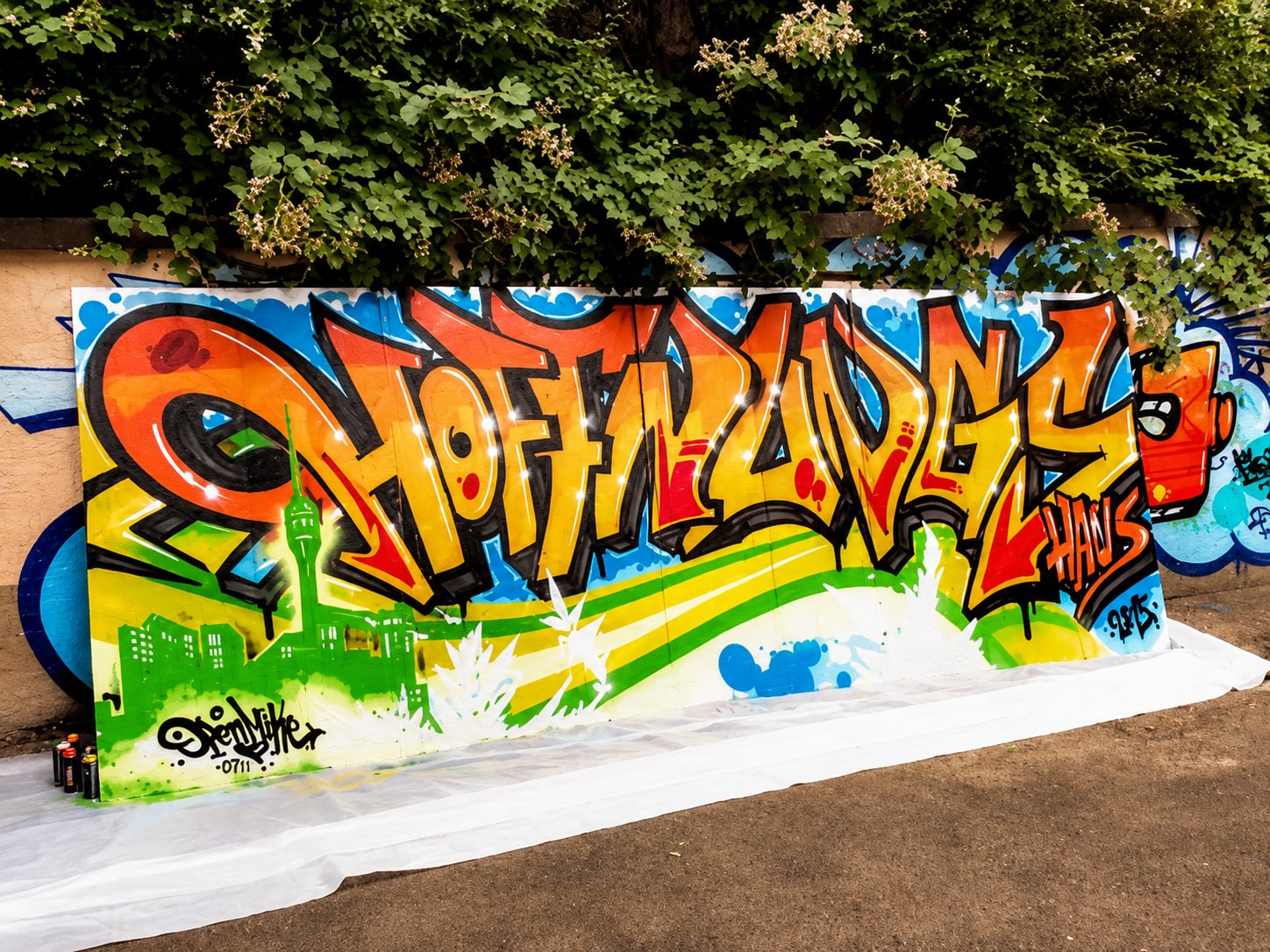 Graffiti-Wandbild mit orange-gelbem Schriftzug 'Hoffnungshaus', darunter grüne Stadtsilhouette mit Kirchturm und Wellenformen auf blauem Hintergrund, vor Hecke aufgestellt, unten Signatur und Spraydosen.