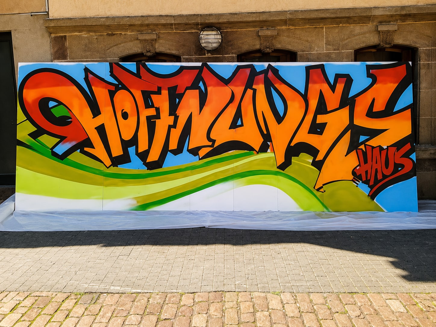 Graffiti-Tafel mit orangefarbenem Schriftzug 'Hoffnungshaus' auf blauem Grund, darunter grün-gelbe Wellenlinien, aufgestellt vor einer Sandsteinfassade mit Rundbogenfenstern.