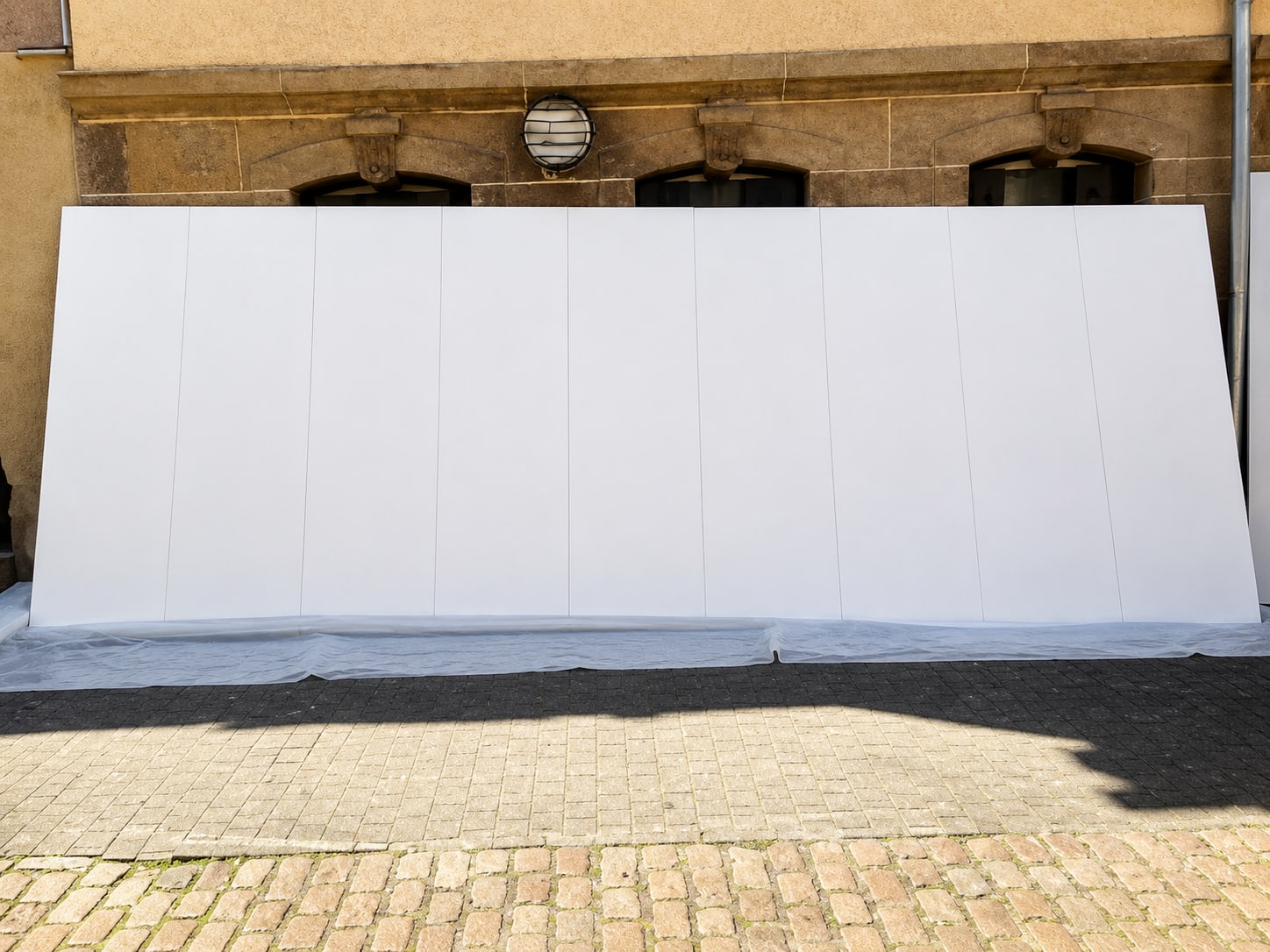Leere weiße Klapptafel aus mehreren Segmenten, aufgestellt vor einer Sandsteinfassade mit drei Rundbogenfenstern und einer Lampe, Untergrund mit Plane abgedeckt.