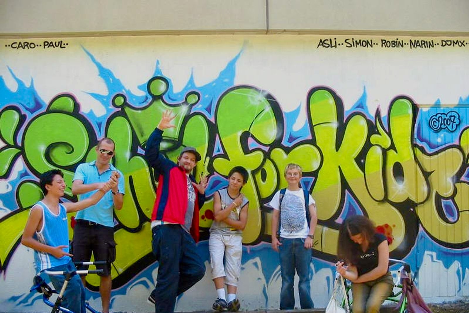 Kinder posieren vor großem Graffiti-Piece mit grünem »Kids«-Schriftzug und blauen Flammen an heller Wand