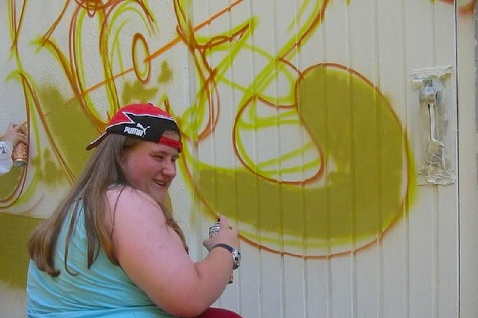 Mädchen mit Basecap sprüht gelb-grüne Graffiti-Outlines an eine weiße Metallwand während des Workshops