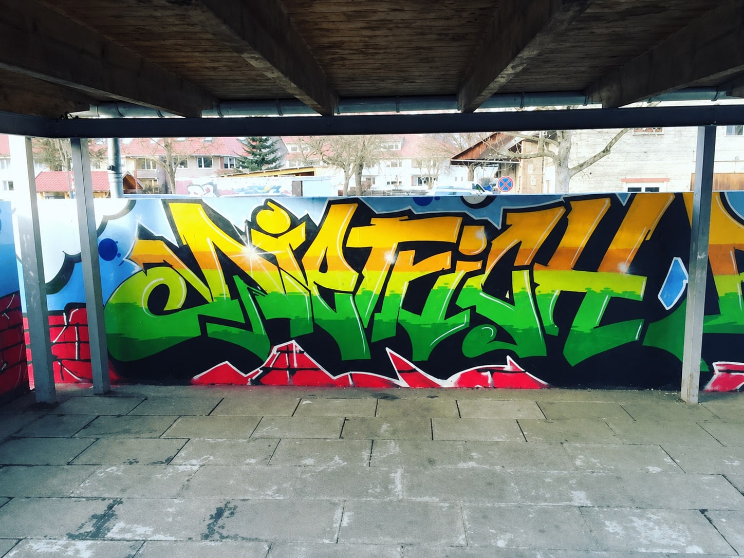 Graffiti-Schriftzug DIETRICH in gelb-grünen Buchstaben mit schwarzer Kontur vor blauem Wolkenhimmel und gemalter roter Ziegelwand, überdachter Gang davor.