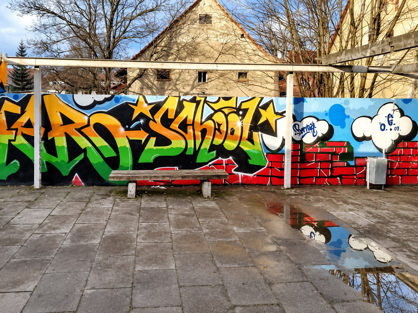 Graffiti-Schriftzug SCHOOL in orange-grünen Buchstaben mit Sternen, daneben eine blaue Wolke mit Signatur und eine gemalte rote Backsteinmauer, davor eine Holzbank auf dem Pausenhof.