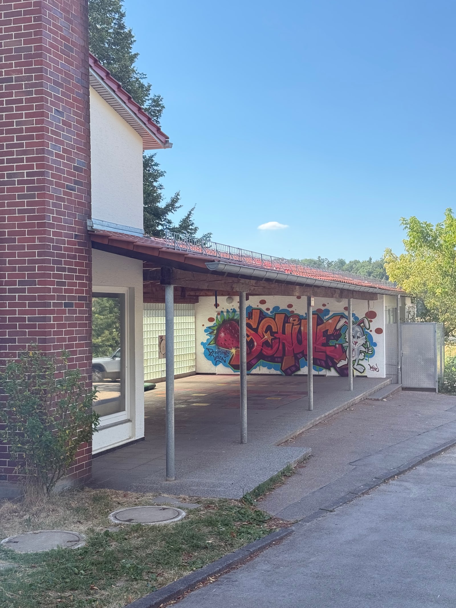 Außenansicht eines überdachten Pausenhof-Unterstands mit Ziegeldach auf Metallstützen, an der Rückwand das bunte Wandgemälde mit Schriftzug 'SCHULE', im Vordergrund ein Backsteingebäude und blauer Himmel.