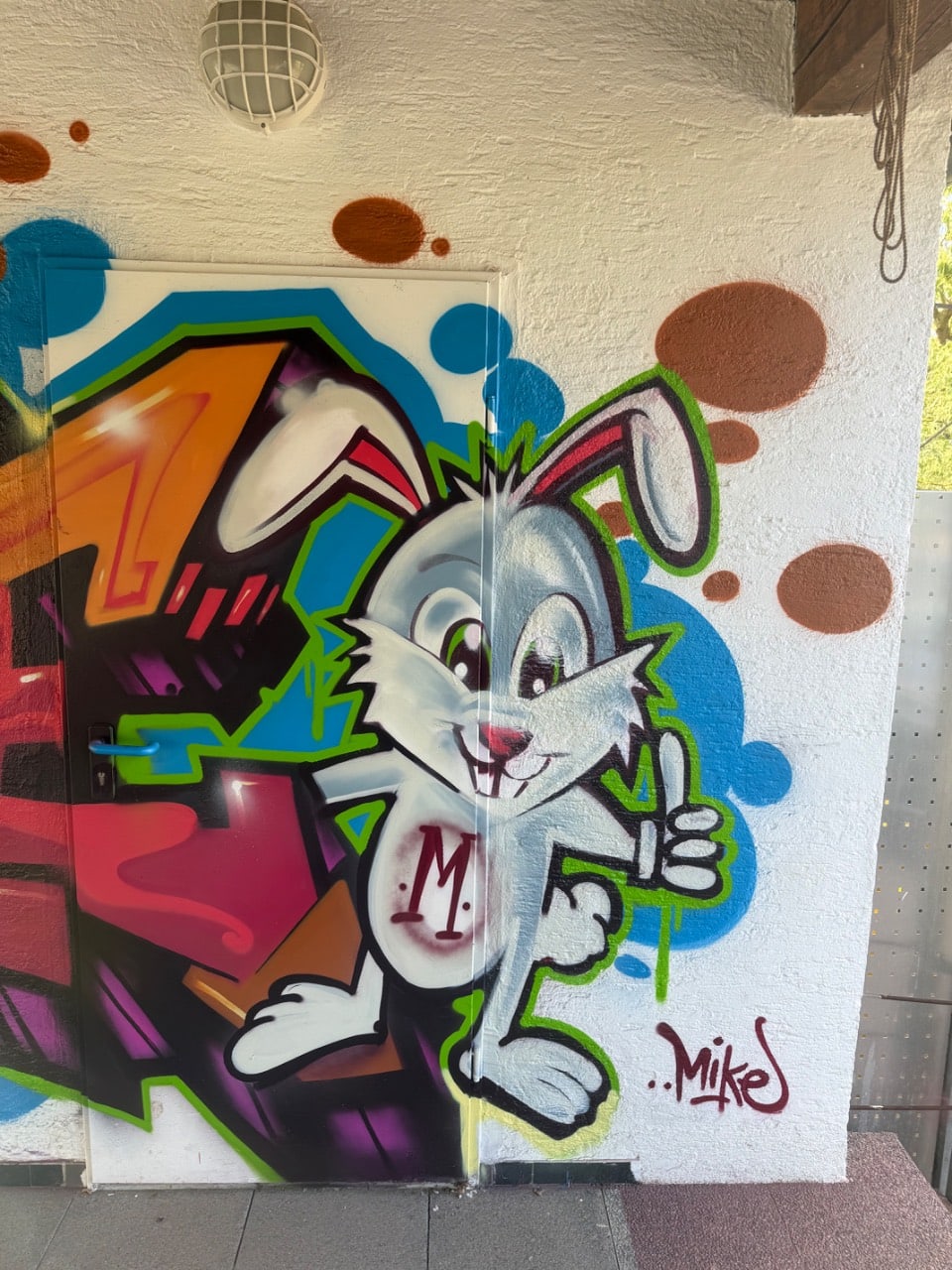Graues Cartoon-Kaninchen mit langen Ohren, rotem Halstuch und 'M'-Zeichen auf der Brust, macht Daumen hoch, daneben Teil eines orangefarbenen Buchstabens, Signatur 'Mike' unten rechts, braune Punkte auf weißer Wand.