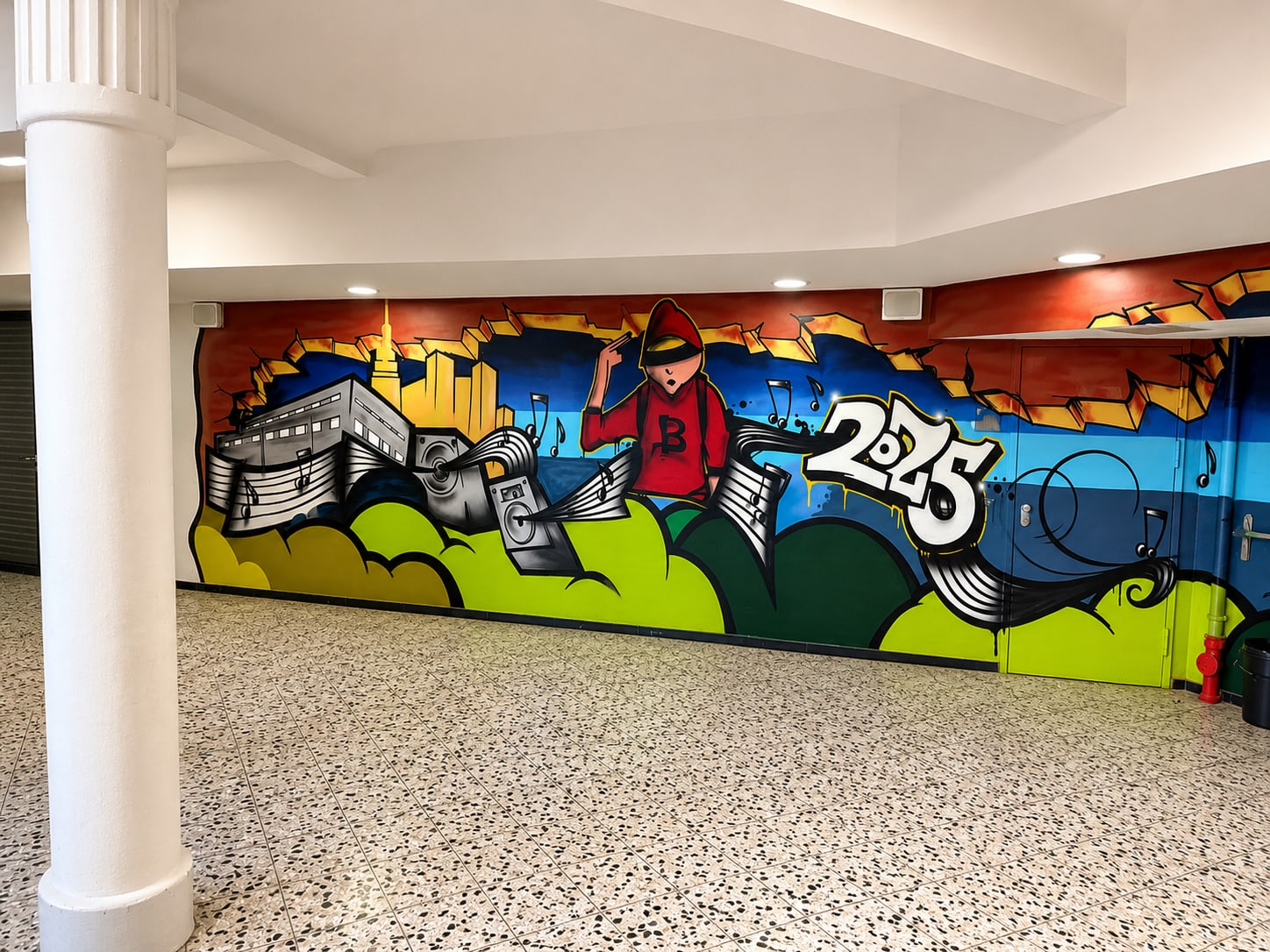 Fertiges Wandbild im Foyer neben weißer Säule: Kapuzenfigur, Lautsprecher, Schriftzug 2025, grüne Hügel, Terrazzoboden