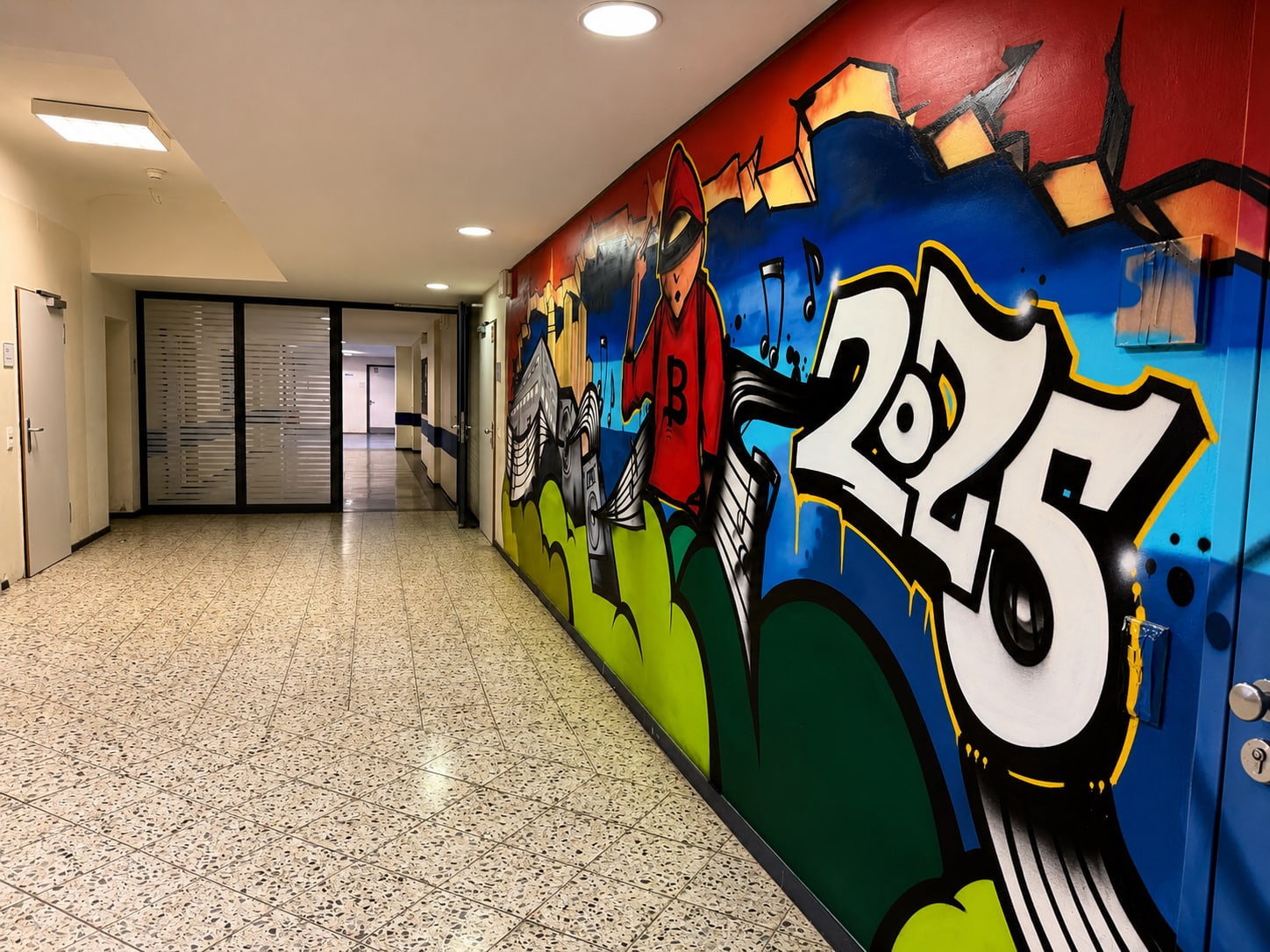 Schulflur mit Terrazzoboden, rechts buntes Wandbild mit Zahl 2025, Lautsprechern, Notenlinien und roter Figur