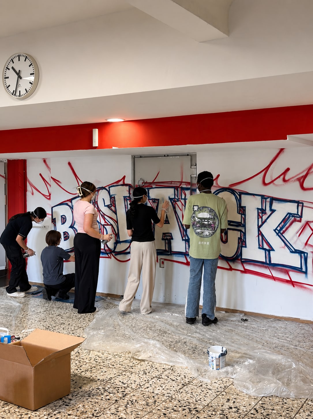 Fünf junge Menschen mit Atemschutzmasken vor weißer Wand mit rot-blauem Umriss-Schriftzug, Folie auf dem Boden