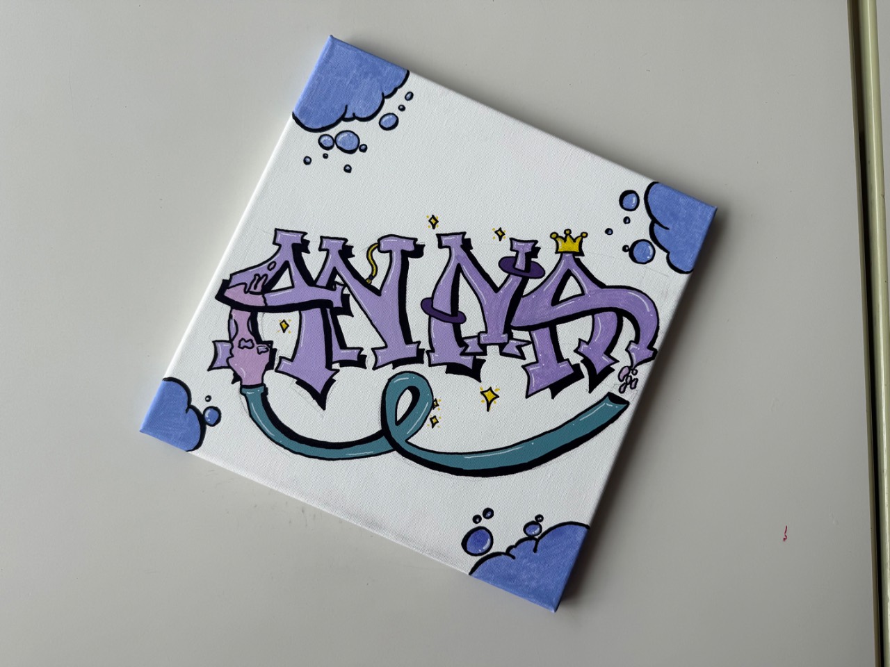 Leinwand mit dem Namen Anna in lila Graffiti-Buchstaben mit kleiner gelber Krone und Glitzersternen, umrahmt von blauen Wolken- und Blasenmotiven in den Ecken.