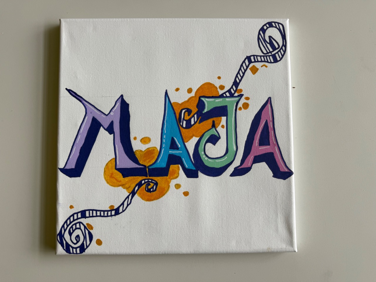 Weiße Leinwand mit dem Namen Maja in bunten Buchstaben (lila, blau, grün, rosa), verziert mit orangen Farbspritzern und einer geschwungenen gestreiften Linie.