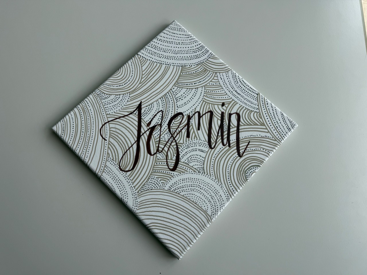 Quadratische Leinwand diagonal auf einem hellgrauen Tisch, bedruckt mit braunen Bogenmustern, darauf in schwarzer Schreibschrift der Name Jasmin.