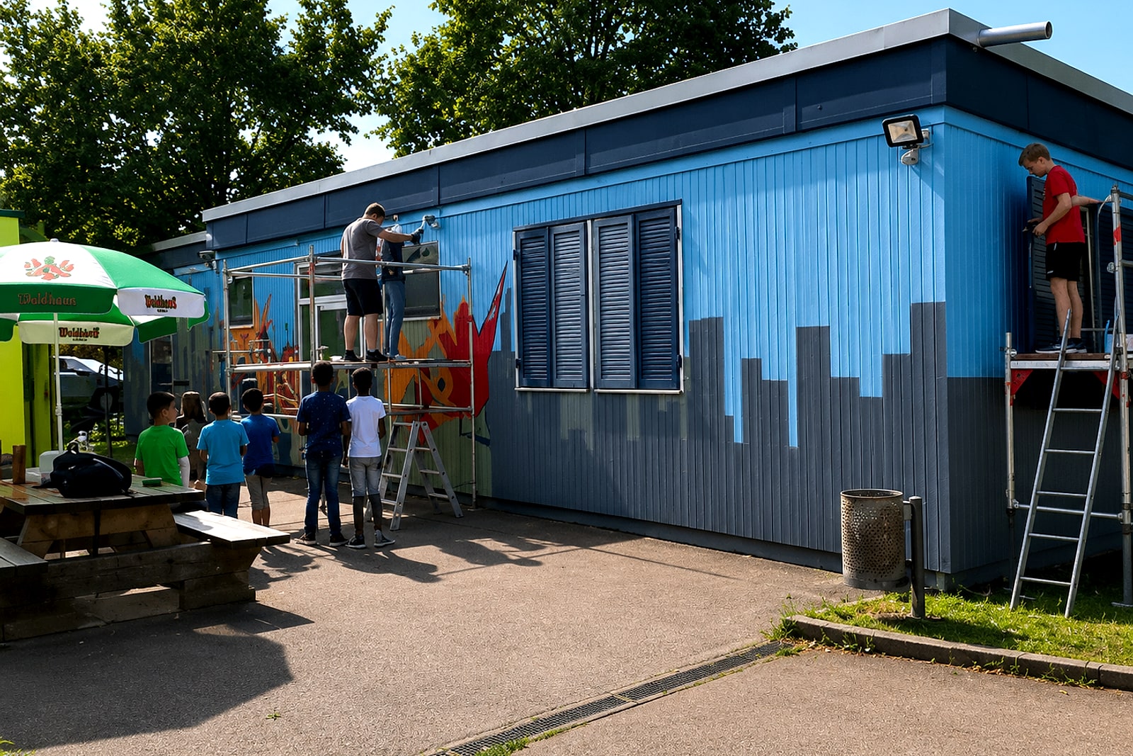Jugendliche gestalten auf einem Gerüst die Fassade des Treffpunkts Rötenberg – Graffiti-Workshop mit Open Mike.