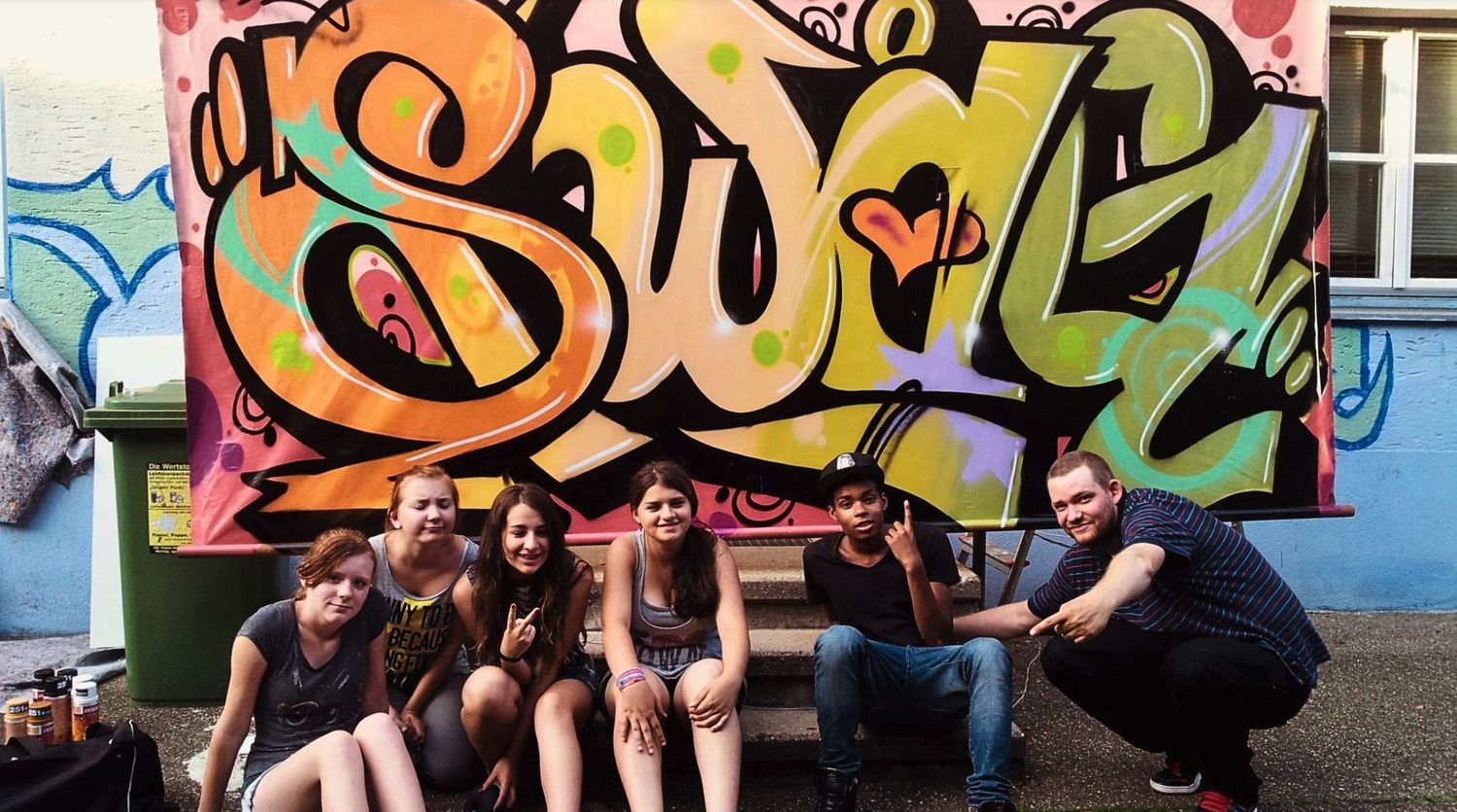 Gruppe Jugendlicher mit Open Mike vor selbst gestaltetem „Swag"-Wildstyle-Mural – Graffiti-Workshop im Jugendhaus Stuttgart